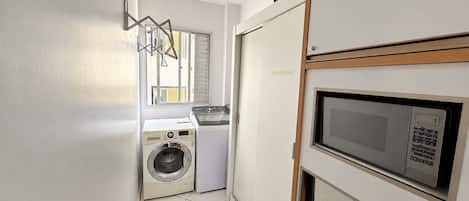 Zona de lavandería con lavadora y secadora, un microondas y una ventana con persianas.