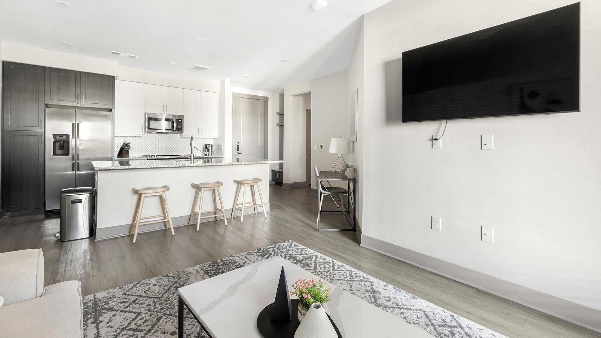 A modern kitchen with stainless steel appliances, a dining area with a table and bar stools, and a mounted flat-screen TV on the wall.