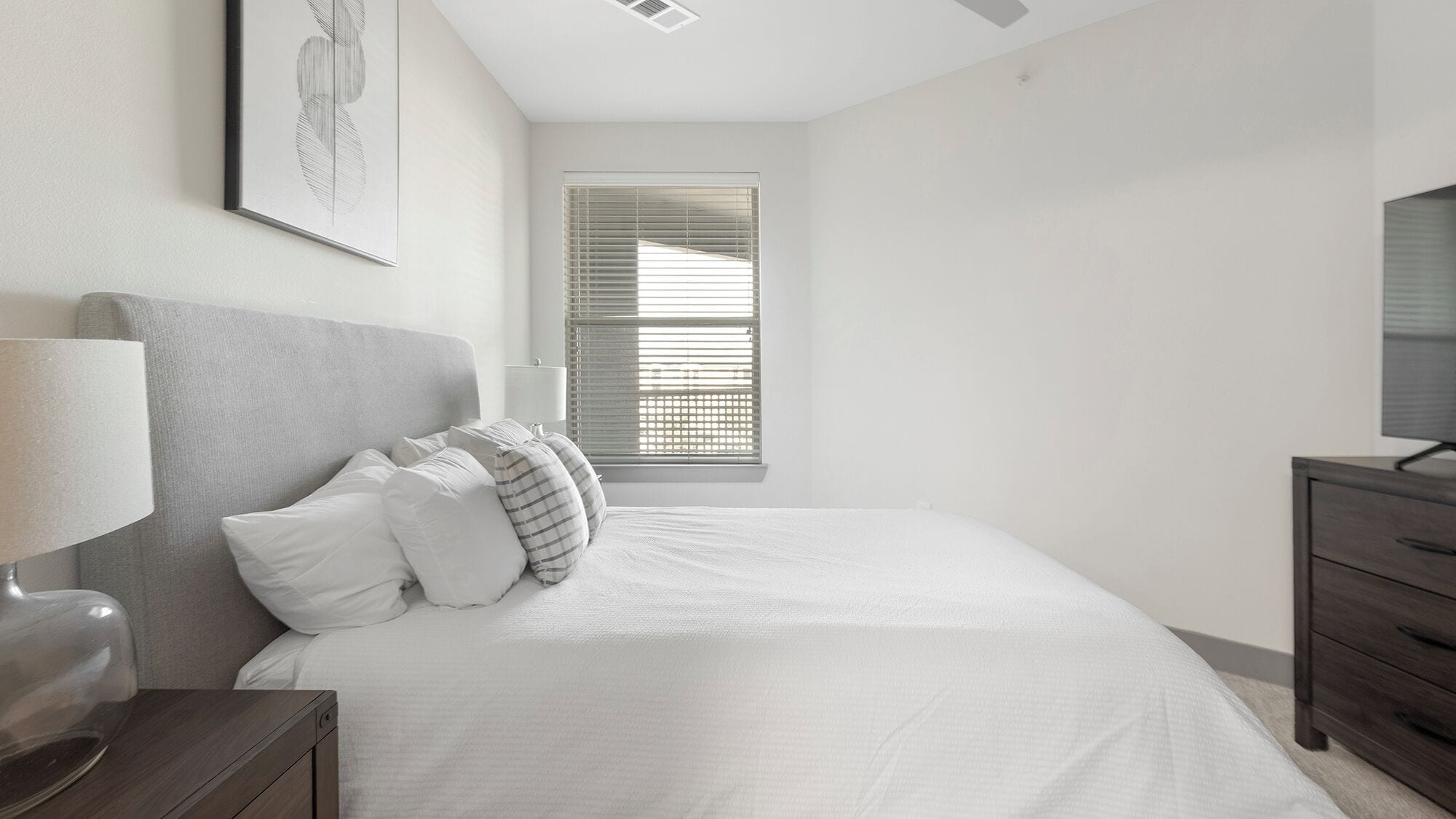 A bedroom with a bed, a nightstand, a lamp, a dresser, and a window with blinds.