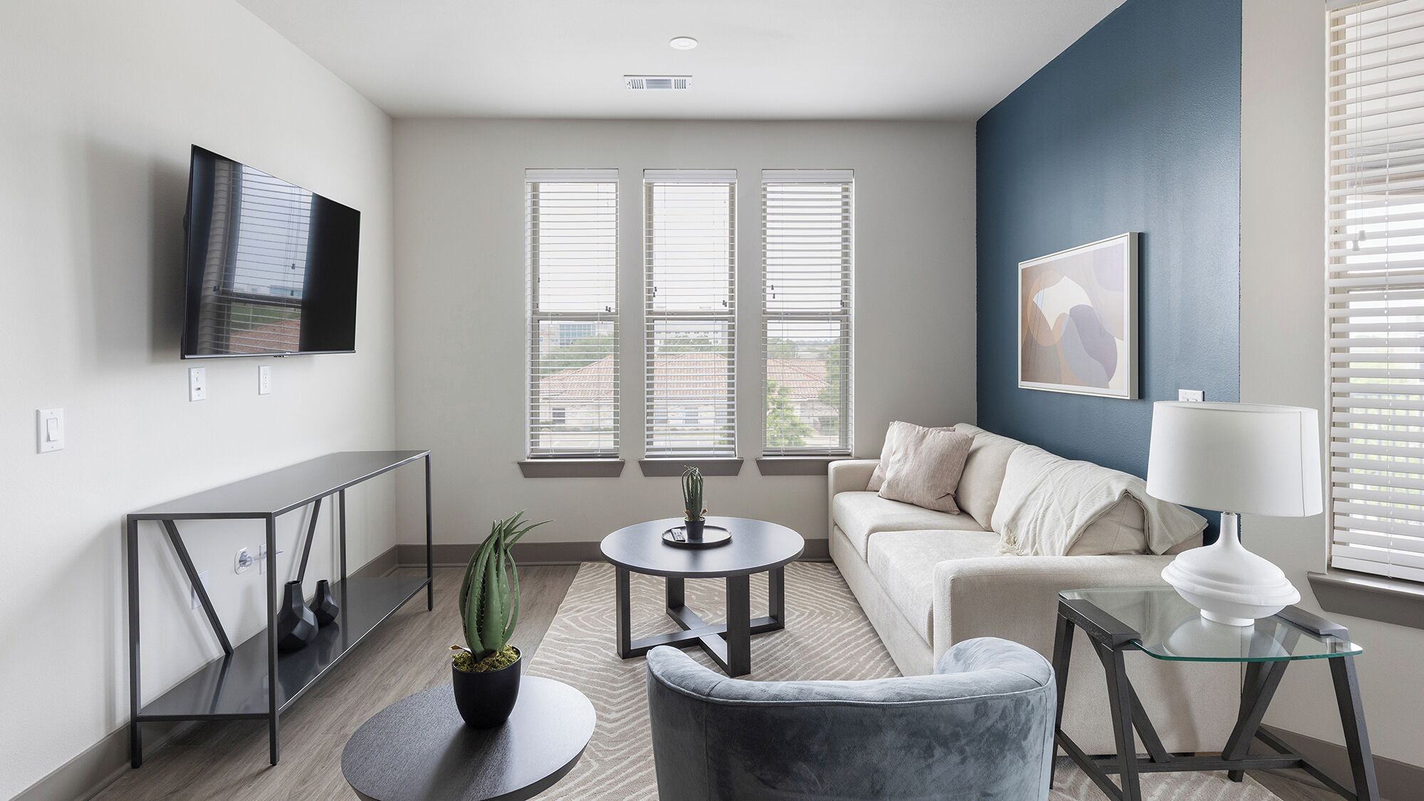 A modern living room with a sofa, a glass coffee table, a TV mounted on the wall, and a window with blinds.