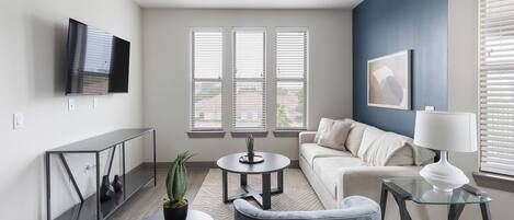 A modern living room with a sofa, a glass coffee table, a TV mounted on the wall, and a window with blinds.