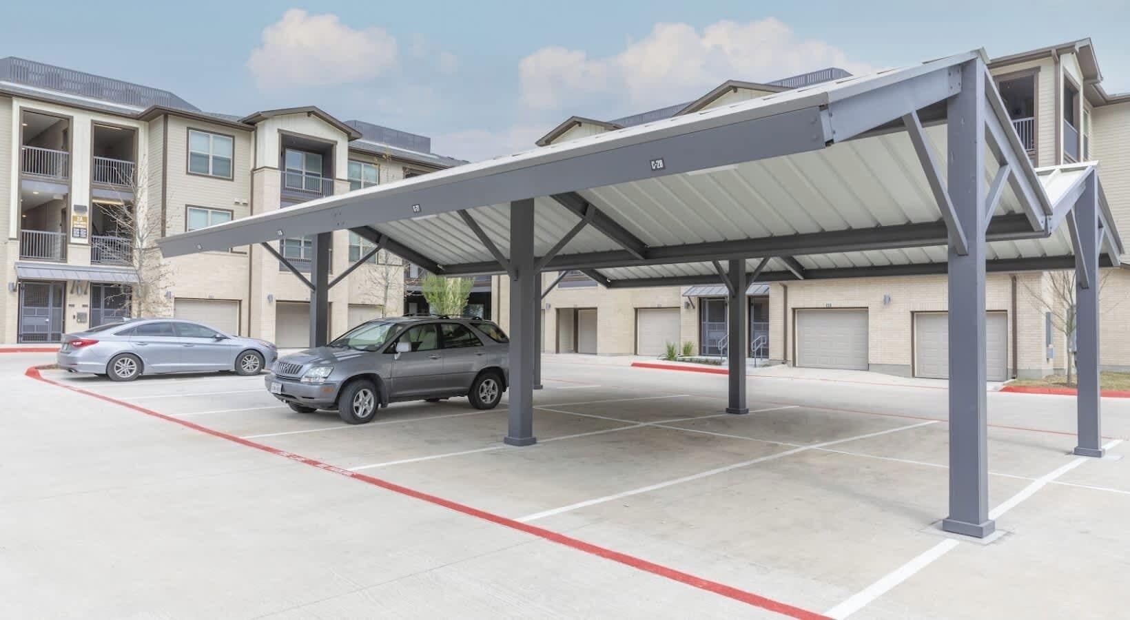 A parking area with a covered structure, a car, and a building with multiple garage doors.