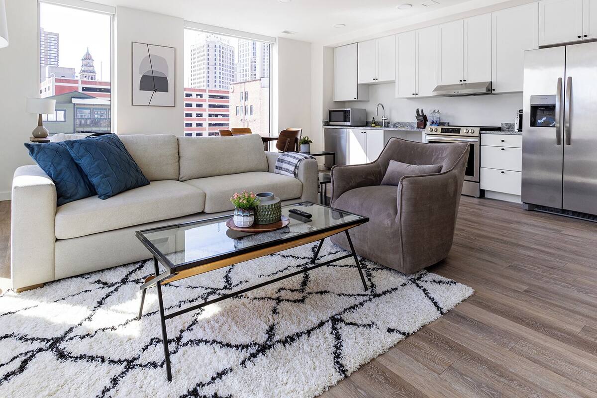 A modern living room with a sofa, armchair, glass coffee table, and a kitchen area with a refrigerator, microwave, and sink.
