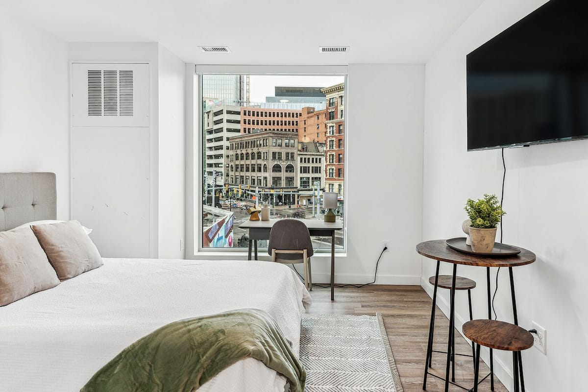 A modern bedroom with a bed, a desk, a chair, a television, and a view of a cityscape through a large window.