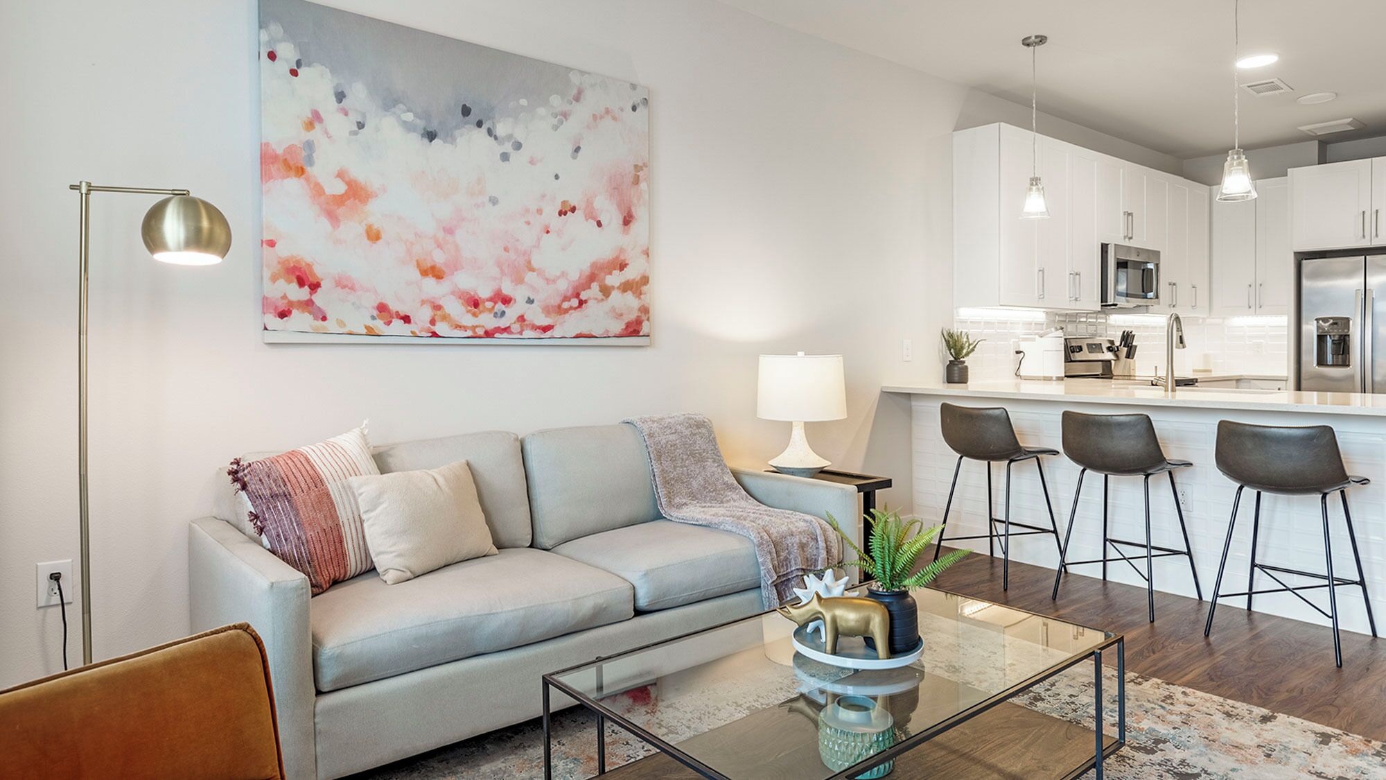 A modern living room with a grey sofa, a glass coffee table, and a kitchen in the background.