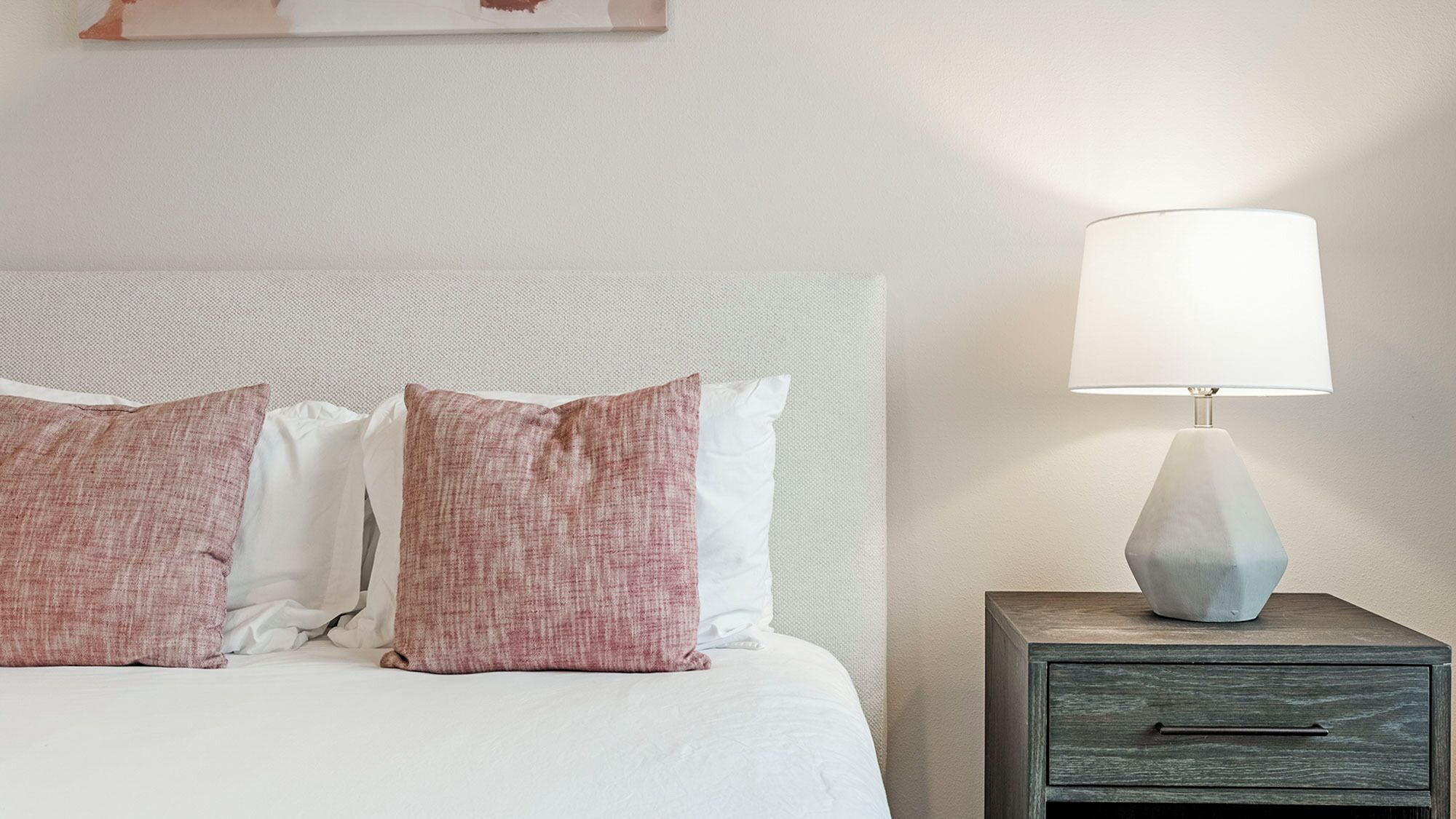 A neatly made bed with white and pink pillows, a modern lamp on a wooden nightstand, and a simple wall with a small artwork.