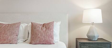 A neatly made bed with white and pink pillows, a modern lamp on a wooden nightstand, and a simple wall with a small artwork.