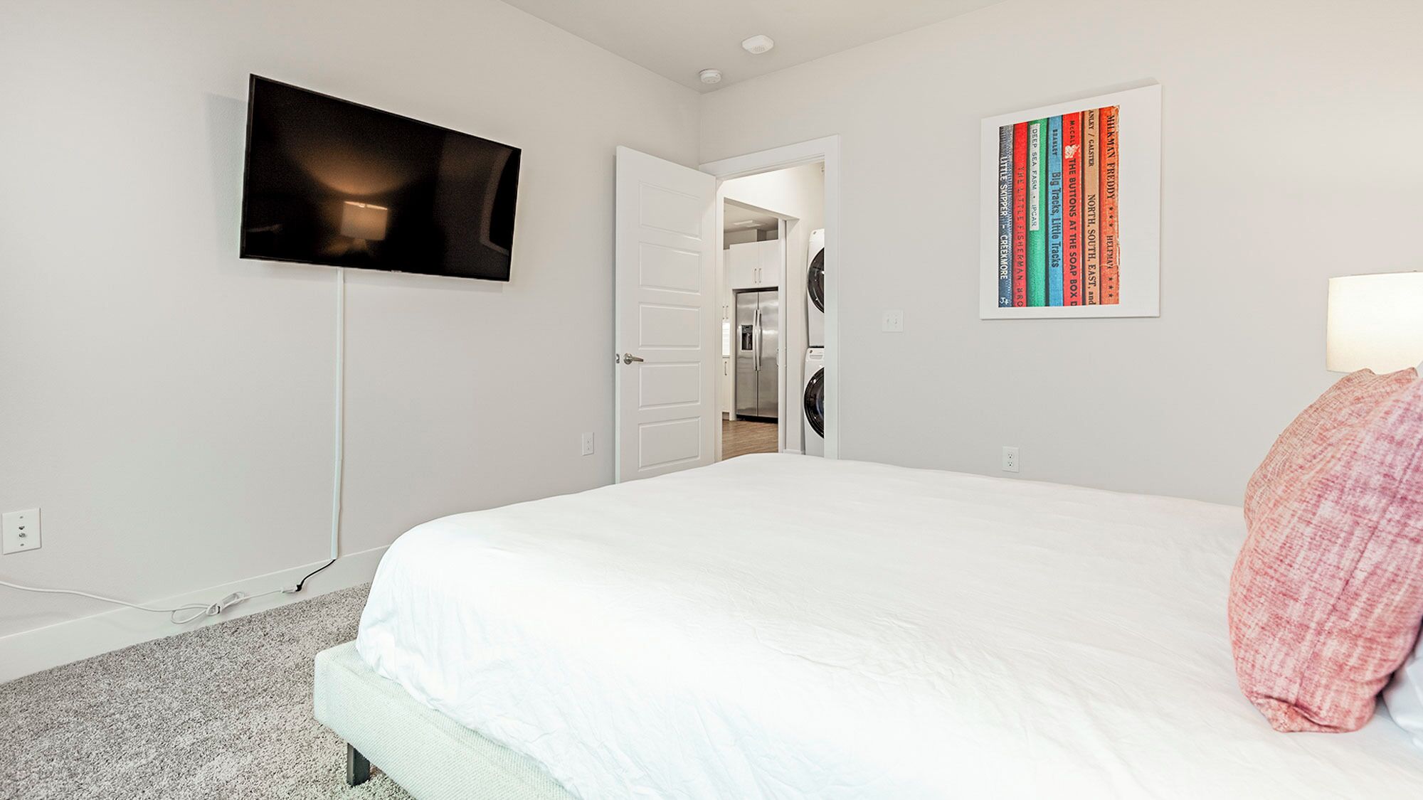 Un dormitorio con cama, televisor montado en la pared, puerta que da a una cocina y una colección de libros enmarcados en la pared.