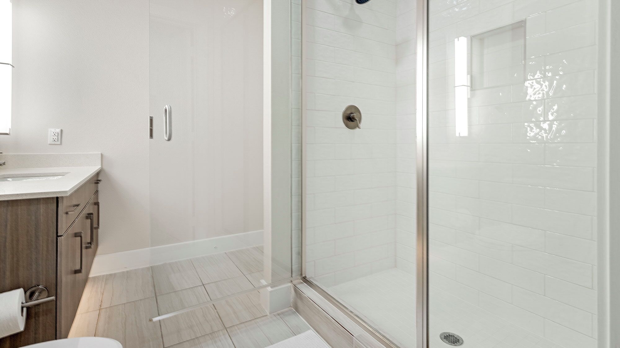 A modern bathroom with a glass shower enclosure, a wooden vanity, and a white tiled floor.