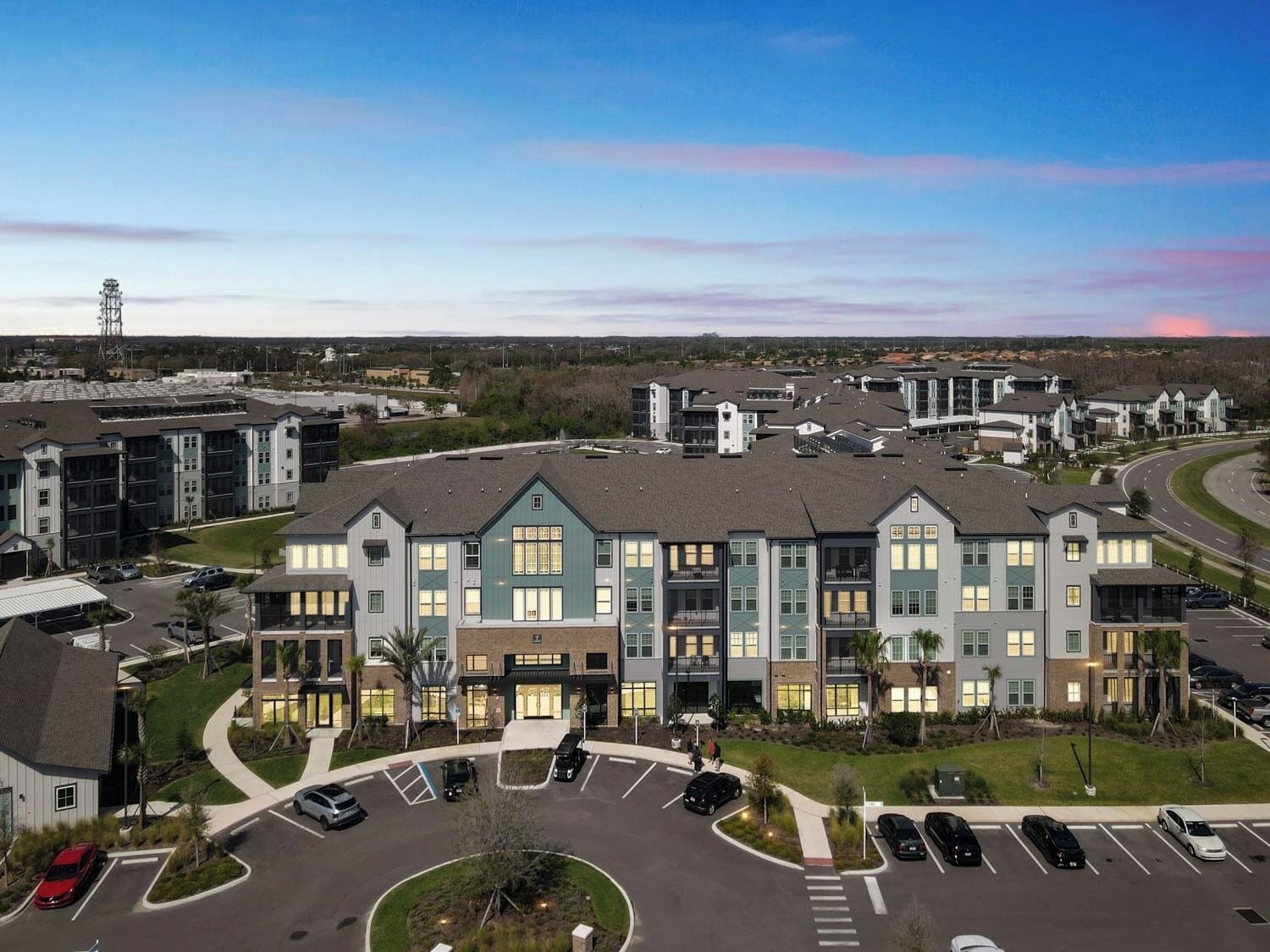 A multi-story residential building with a central courtyard, parking lot, and a road in the background.