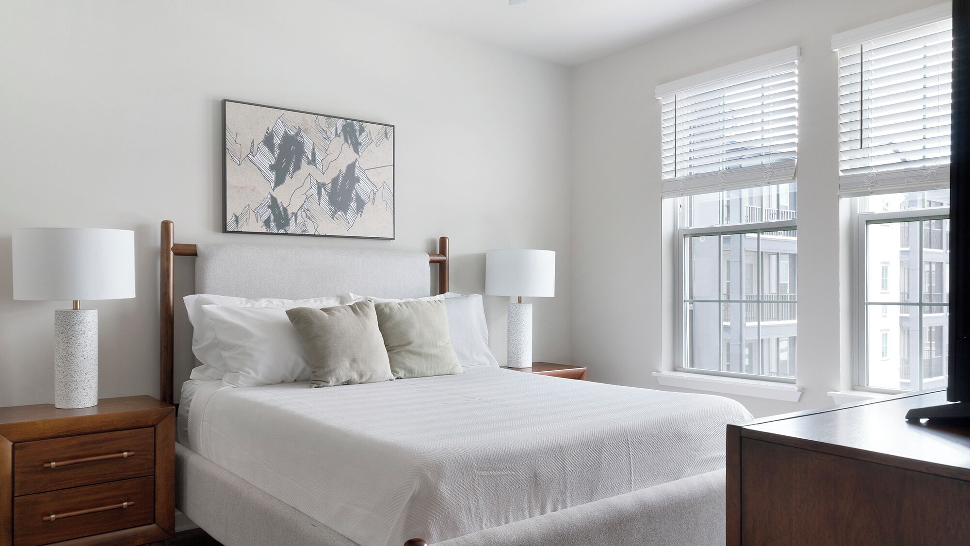 A bedroom with a bed, two bedside tables, a nightstand, a window with blinds, and a wall-mounted artwork.