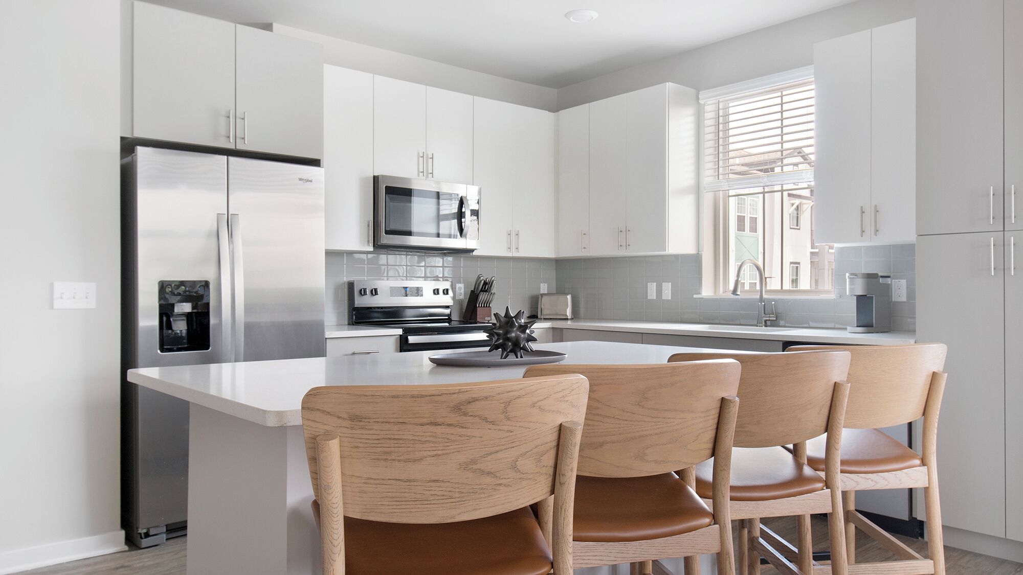 Cocina moderna con armarios blancos, electrodomésticos de acero inoxidable, una isla central con sillas y un ventanal que proporciona luz natural.