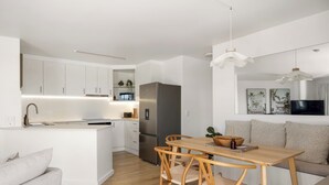 A modern kitchen with white cabinetry, a stainless steel refrigerator, and a dining area with a wooden table and chairs. - Absolute Beachfront, Panoramic Ocean Views & Whale Season Magic (May–August) 🐳 (Coolum Beach)