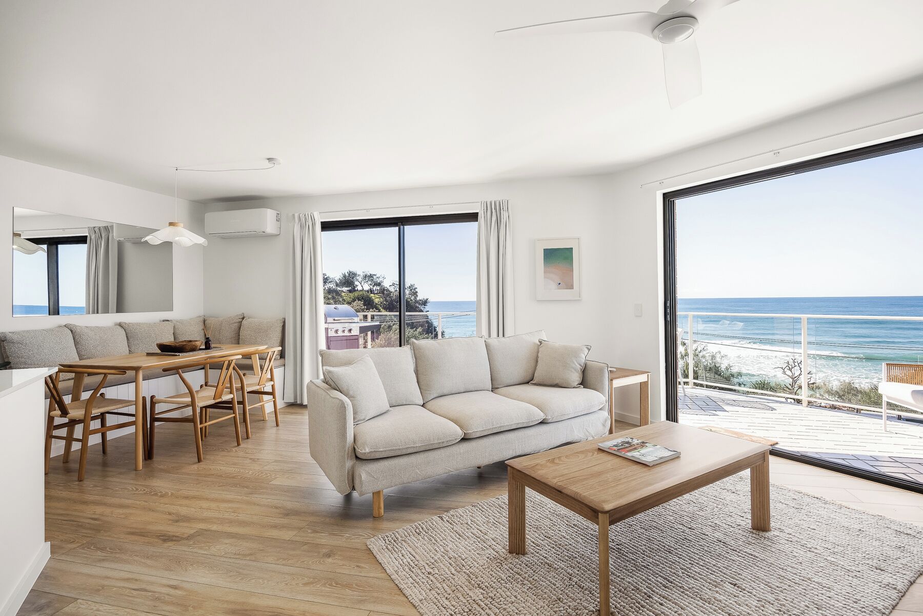 Un soggiorno moderno con tavolo da pranzo in legno, divano beige e tavolino da caffè in legno. È presente una grande finestra con vista sull'oceano e un balcone con ringhiera.