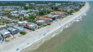 Ein Luftbild einer Küstenstadt mit Strandhäusern, darunter ein Hotel, das mit einem roten Pfeil gekennzeichnet ist, umgeben von viel Grün und einem Sandstrand.