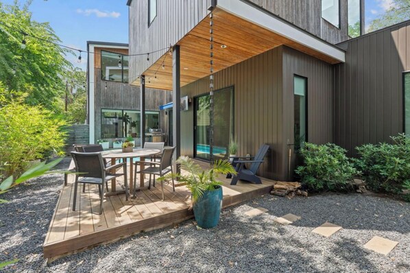 Una casa moderna con un patio que tiene una terraza de madera, mobiliario exterior y una planta grande en maceta.