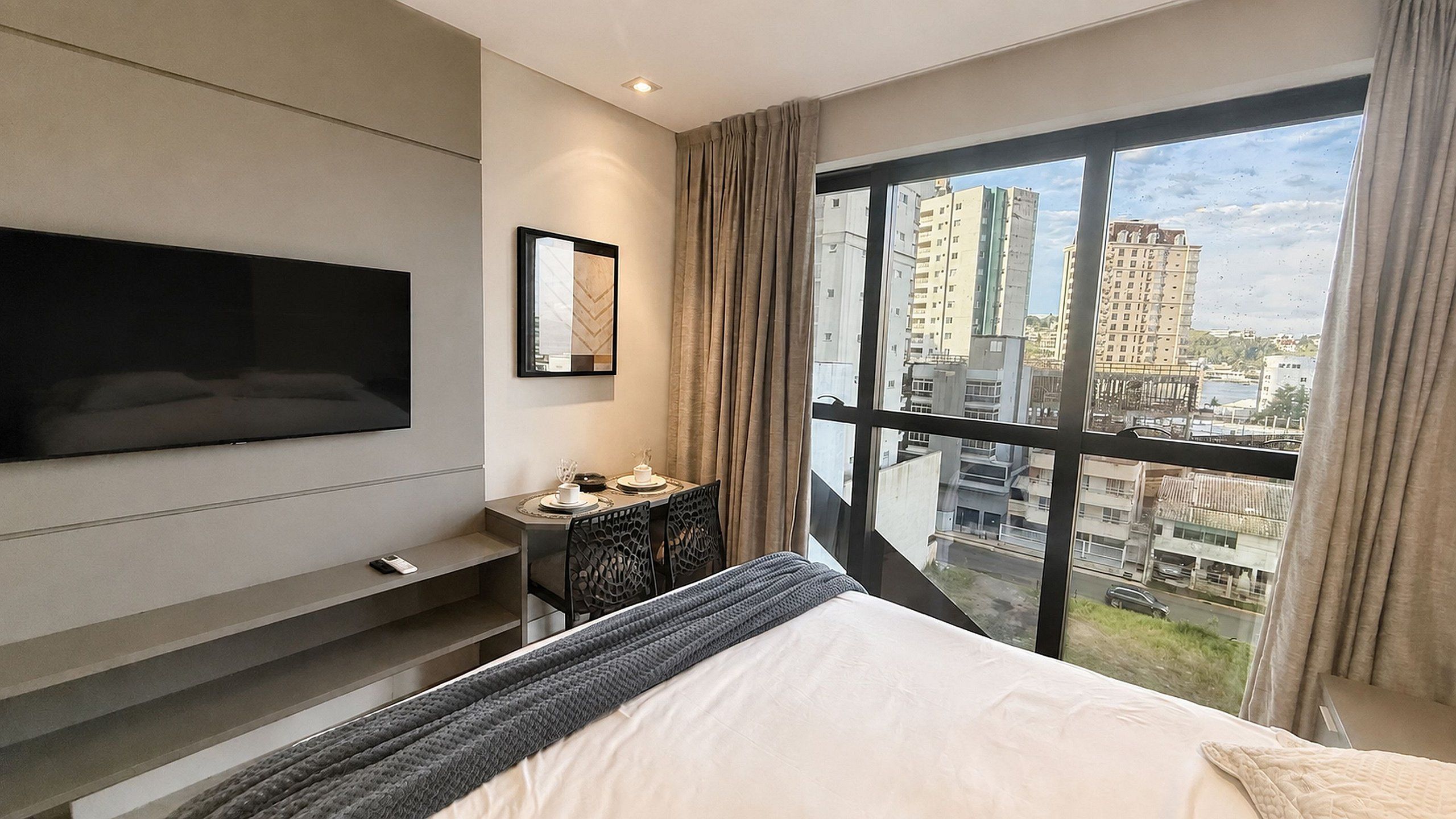 Une chambre d’hôtel moderne dotée d’un grand lit, d’un téléviseur à écran plat fixé au mur, d’une petite table agrémentée d’un objet décoratif et d’une grande fenêtre offrant une vue sur la ville.