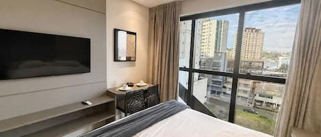 Una habitación de hotel moderna con una cama grande, un televisor de pantalla plana montado en la pared, una mesita con un adorno y un ventanal que ofrece vistas a la ciudad.