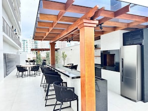 An outdoor kitchen area with a stainless steel refrigerator, wooden pergola, and black chairs. - Manhattan Flats (Itapema)