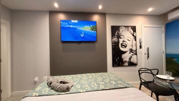 Habitación de hotel con cama, televisión, un cuadro enmarcado, una silla y una mesa con un vaso.