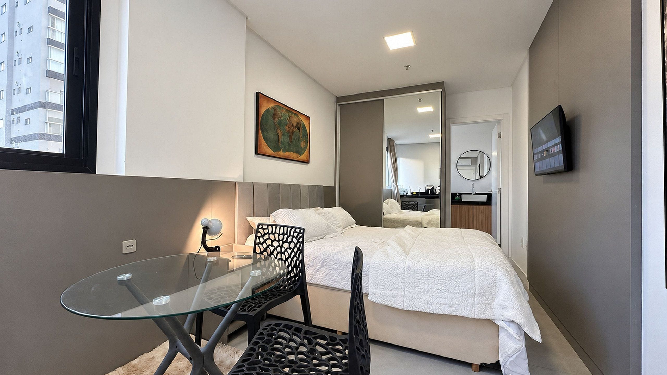 Habitación de hotel pequeña con una cama, una mesa de vidrio, una silla, un televisor y un cuadro en la pared.