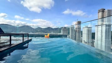 A rooftop infinity pool with a glass barrier, a wooden deck, and a view of surrounding high-rise buildings and mountains.