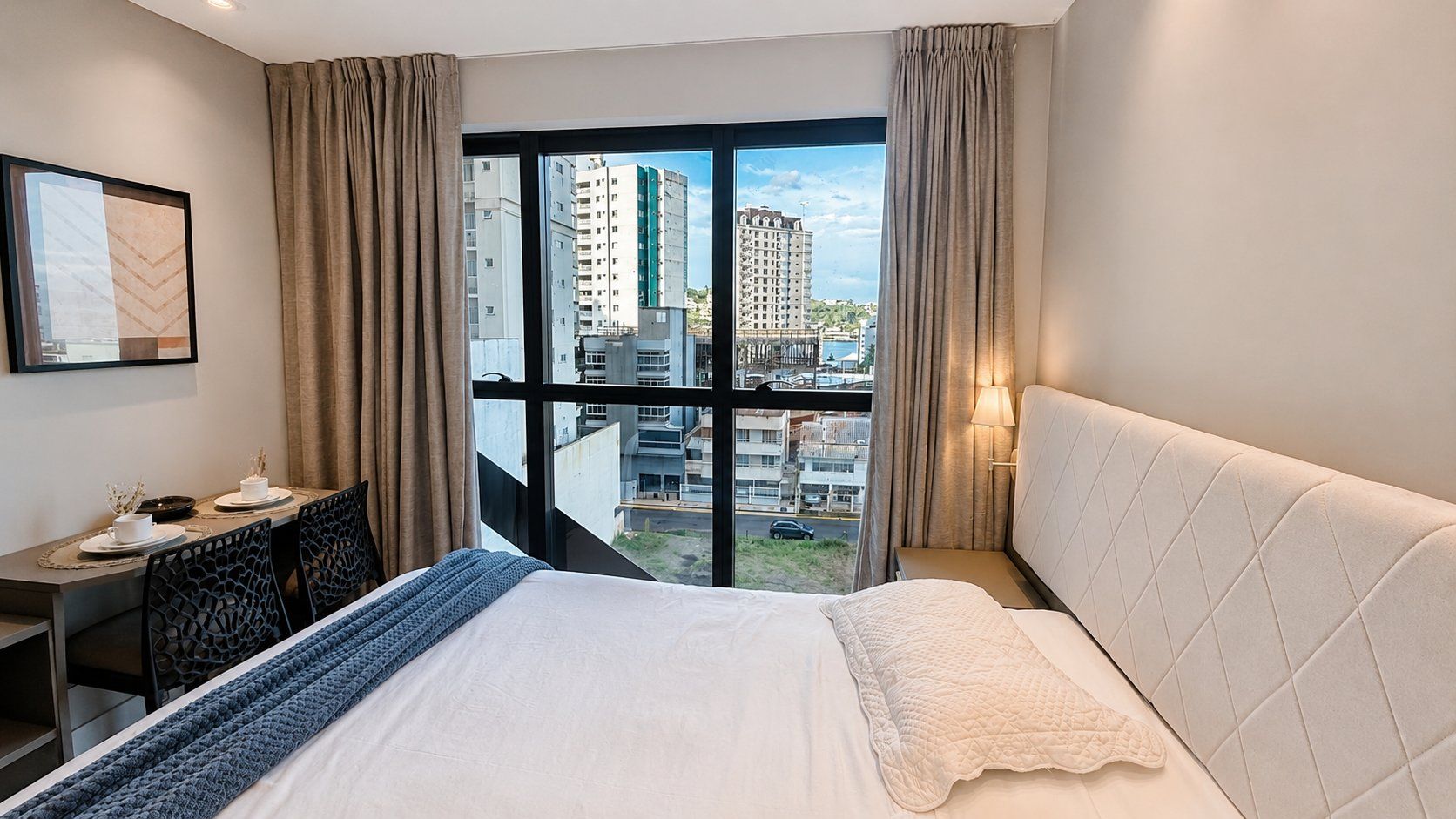 A hotel room with a large bed, a desk, and a view of the cityscape through a large window.