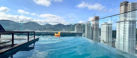Ein Dachpool mit unendlicher Optik, einer Glaskante, einer Holzterrasse und Blick auf die umliegenden Hochhäuser und Berge.