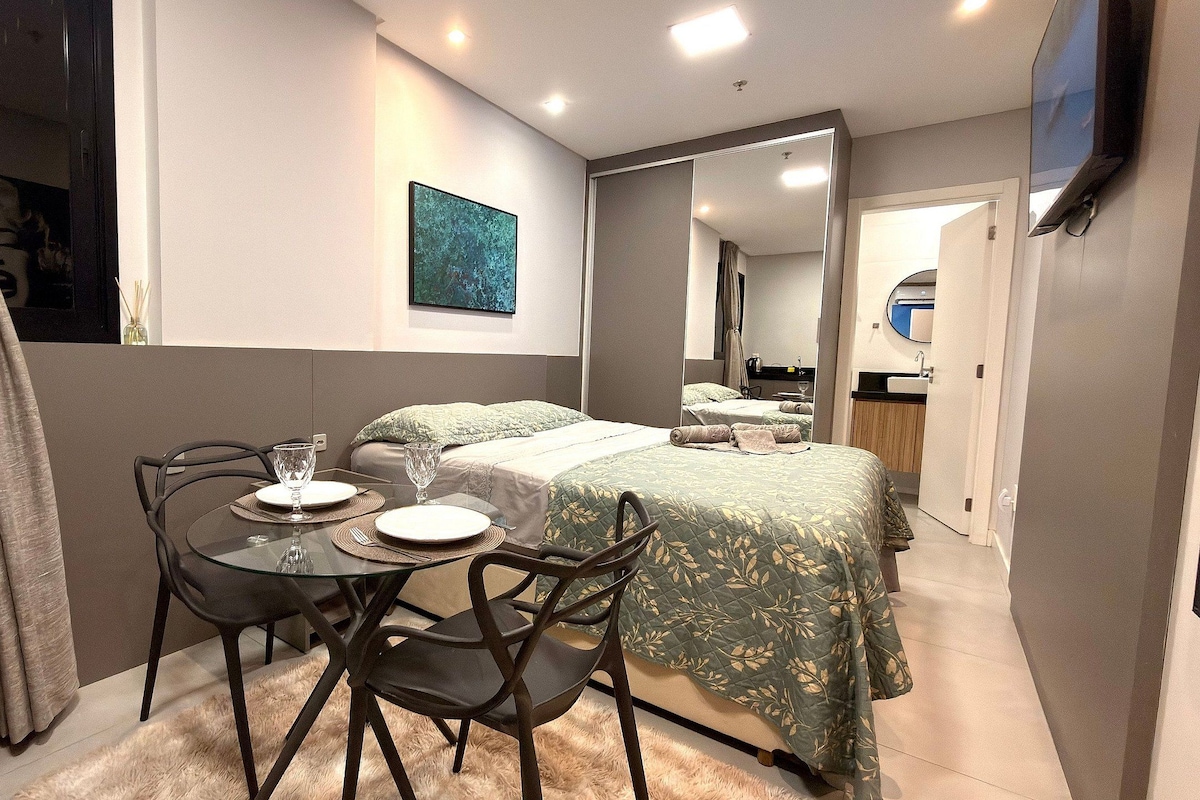 A compact hotel room with a bed, a small dining table with chairs, a mirror, and a television mounted on the wall.