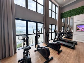 A gym with various exercise equipment, large windows, and a view of a cityscape. - Manhattan Flats (Itapema)