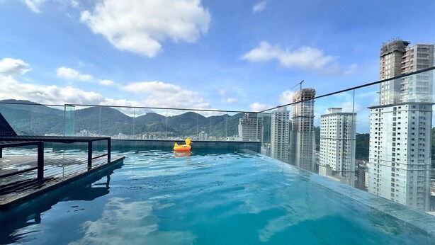 Piscina infinita no terraço, com barreira de vidro, deck de madeira e vista para edifícios altos e montanhas ao redor.