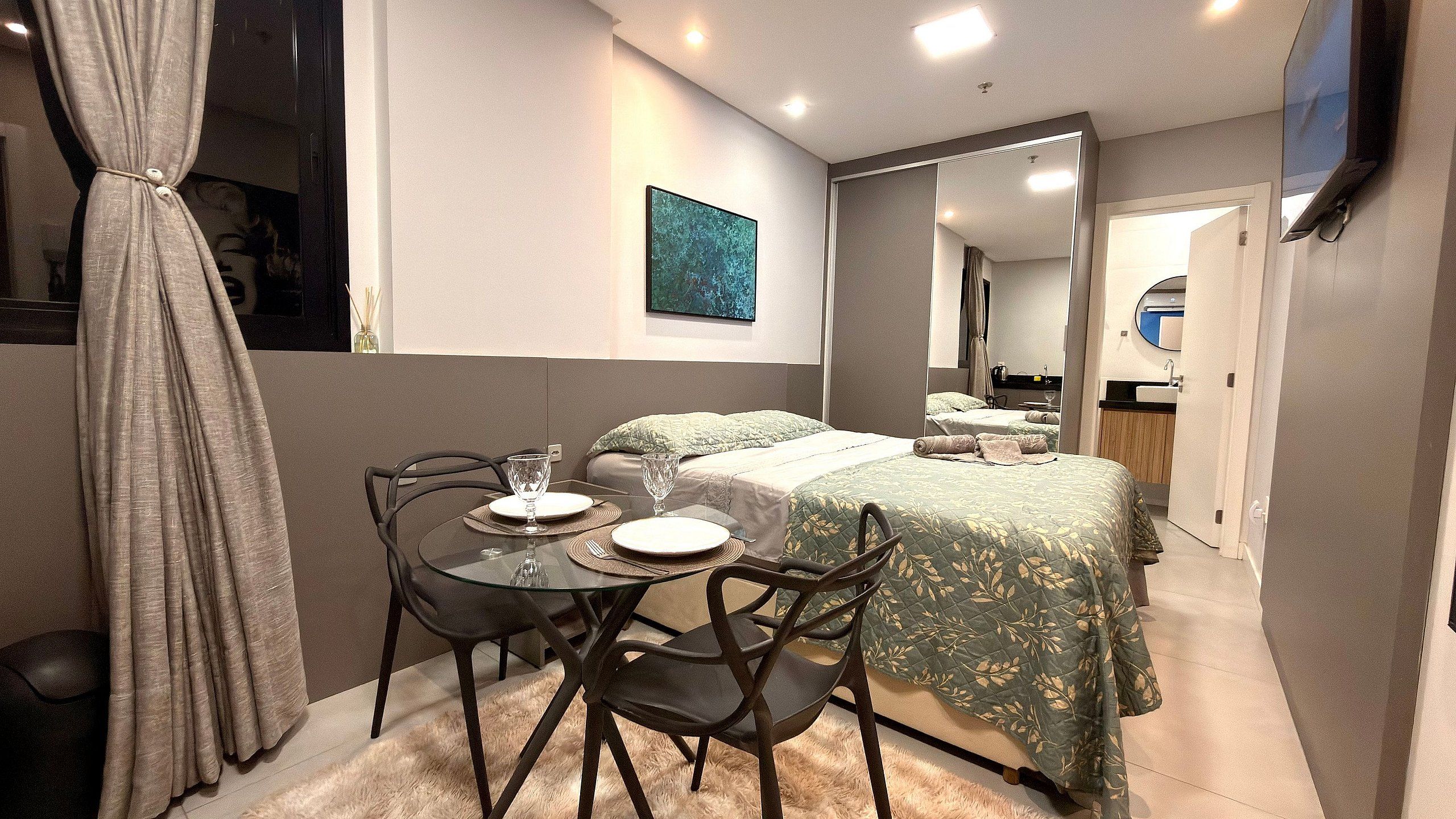 A compact hotel room with a bed, a small dining table with chairs, a mirror, and a television mounted on the wall.
