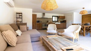 Una sala de estar moderna con un sofá, sillones y una mesa de centro. Se ve una cocina al fondo con armarios de madera y un comedor con una mesa y sillas.