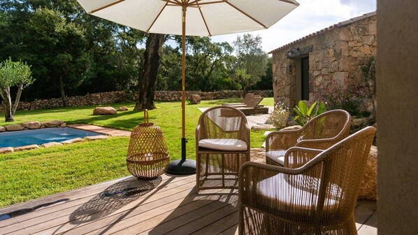 A deck with wicker furniture and a large umbrella, overlooking a swimming pool and a stone building.