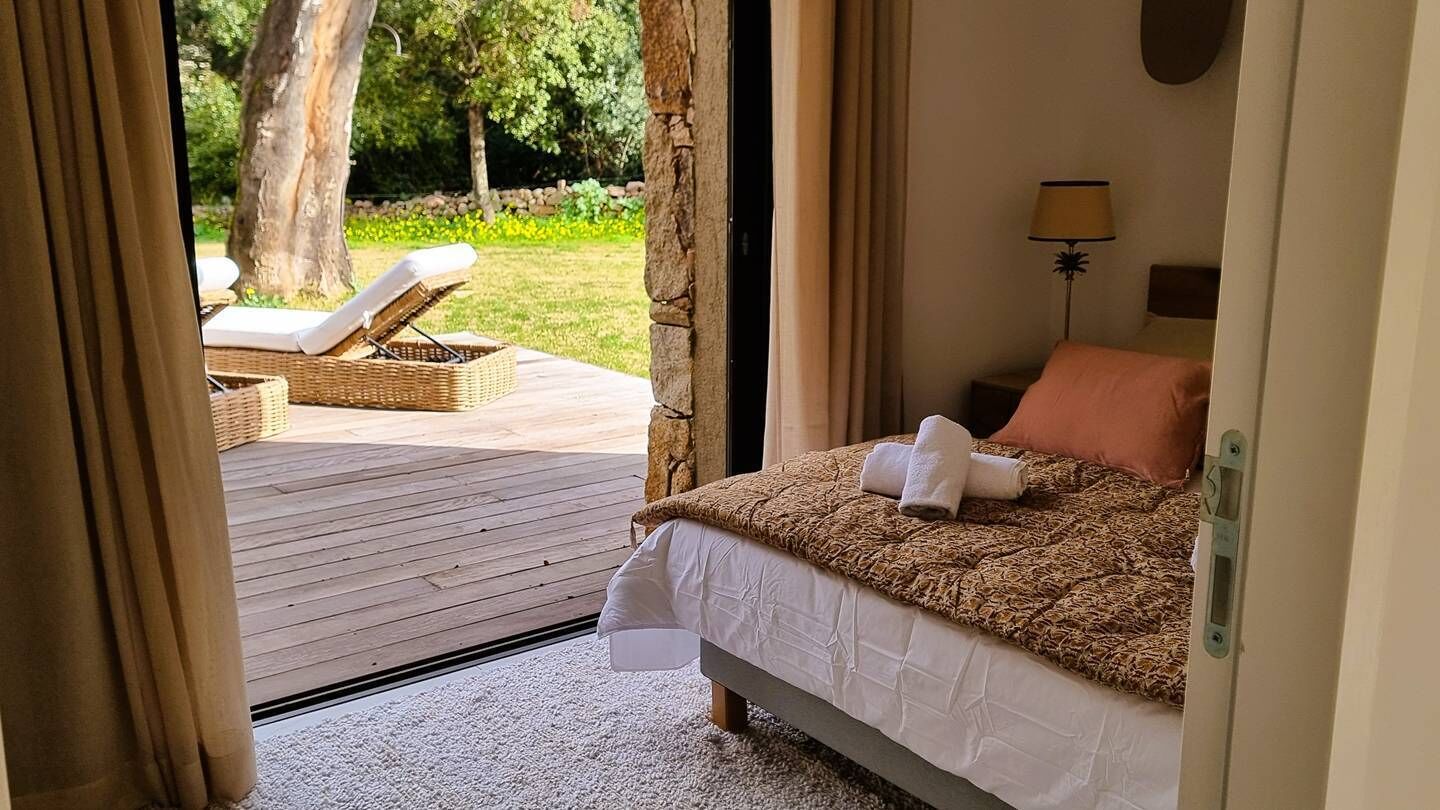 A bedroom with a bed, a nightstand, a lamp, and a view of an outdoor deck with a chair and a table.