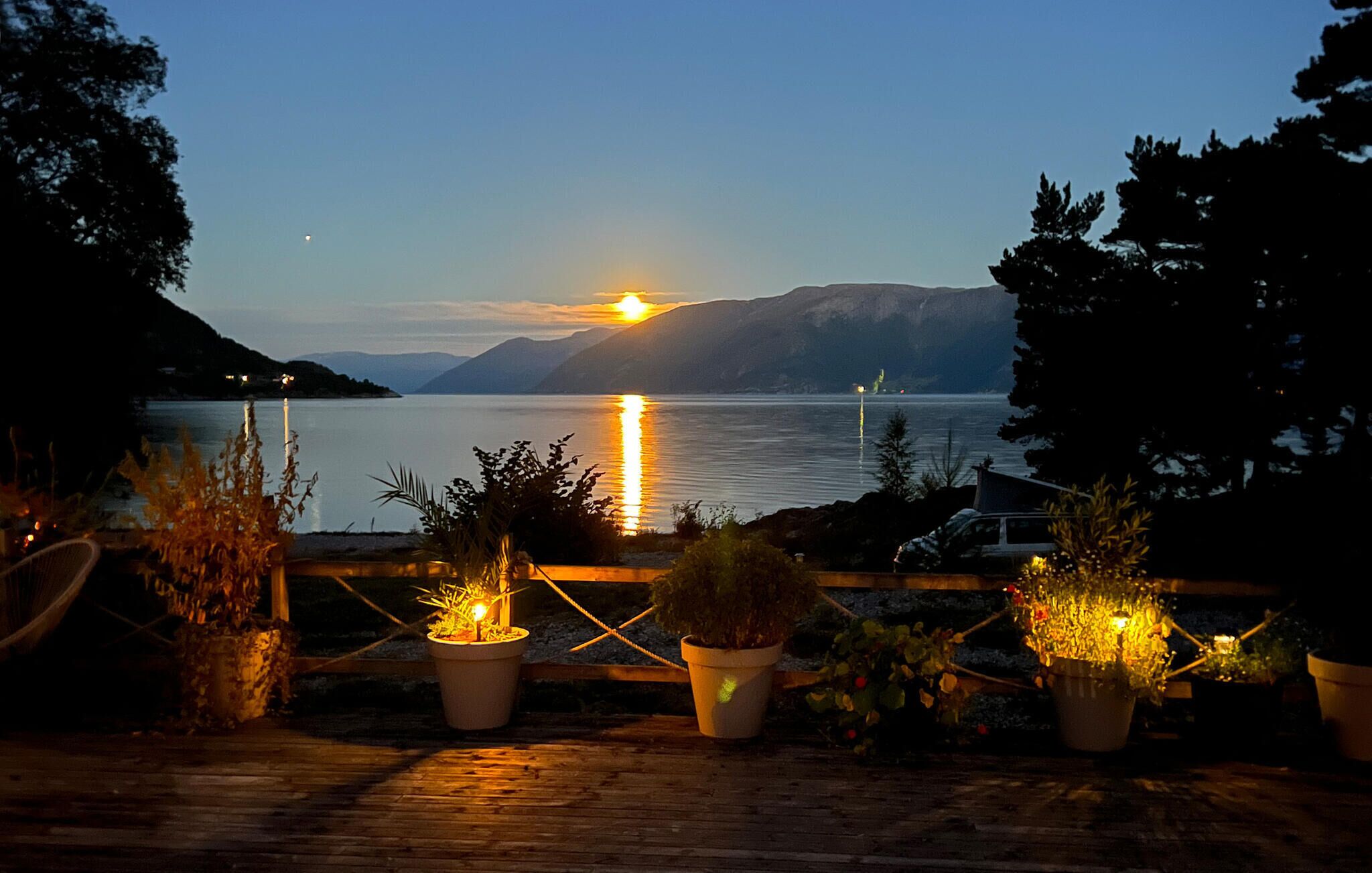 Vista de um lago ao pôr do sol, com montanhas ao fundo e plantas em vasos no primeiro plano.