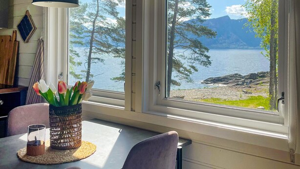 Ein Essbereich mit Tisch, Stühlen, einer Vase mit Blumen und Blick auf einen See mit Bäumen und Bergen.