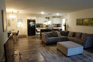 A modern living room with a gray sectional sofa, a white ottoman, and a patterned rug. There is a kitchen area with white cabinets and a black refrigerator. - Meadow Lane In Hocking Hills (Logan)