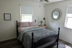 Une chambre à coucher avec un lit, une fenêtre avec un store blanc, un miroir fixé au mur, un tableau encadré et une petite table avec une lampe.