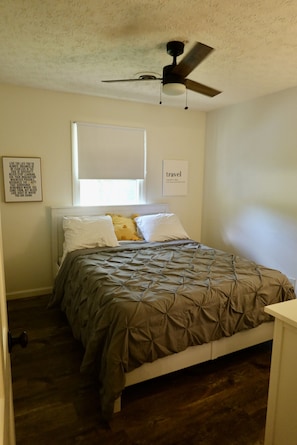 Quarto com cama, ventilador de teto, janela com persianas e duas molduras com citações na parede.