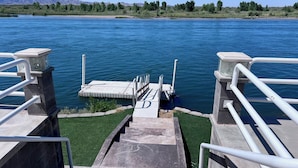 Una escalera de concreto que lleva a un muelle con barandilla de metal, rodeado de agua y una cadena montañosa en la distancia.