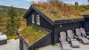 A black building with a sloped roof, a small porch with Adirondack chairs, and a grassy hill in the background. - Cozy home in Hovden i Setesdal - with jacuzzi (Hovden i Setesdal)
