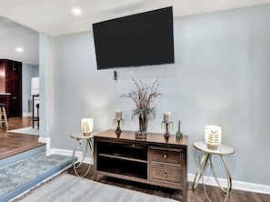 A modern living room with a flat-screen TV mounted on the wall, a dark wooden console table, two small round tables with decorative lamps, and a patterned rug.