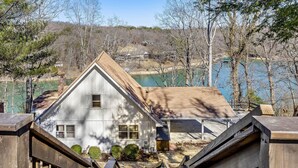 A white house with a steep roof, wooden deck, and a view of a lake and hills in the background.