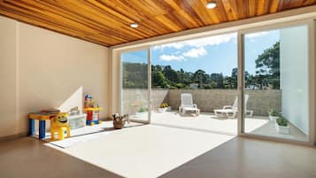 Una habitación amplia con techo de madera, un gran ventanal corredero que da a un patio exterior con sillas blancas y plantas en macetas.