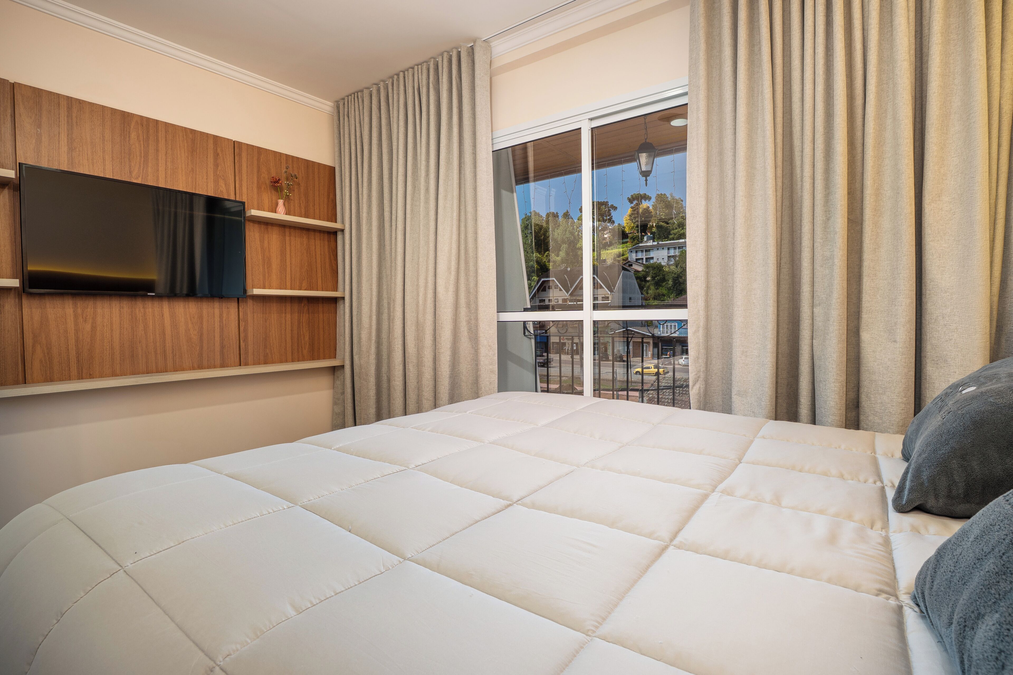 Quarto com cama grande, televisão montada na parede e janela com cortinas.