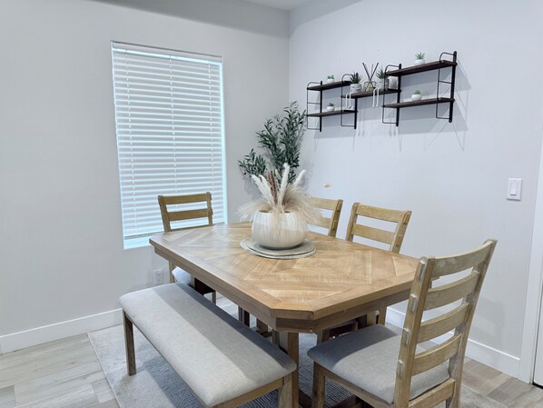 A dining area with a wooden table, four chairs, a bench, and a potted plant. There are three shelves on the wall above the table.