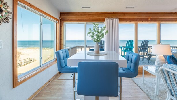 Una habitación con un amplio ventanal con vistas a la playa, una mesa blanca y sillas azules, y vista al océano.