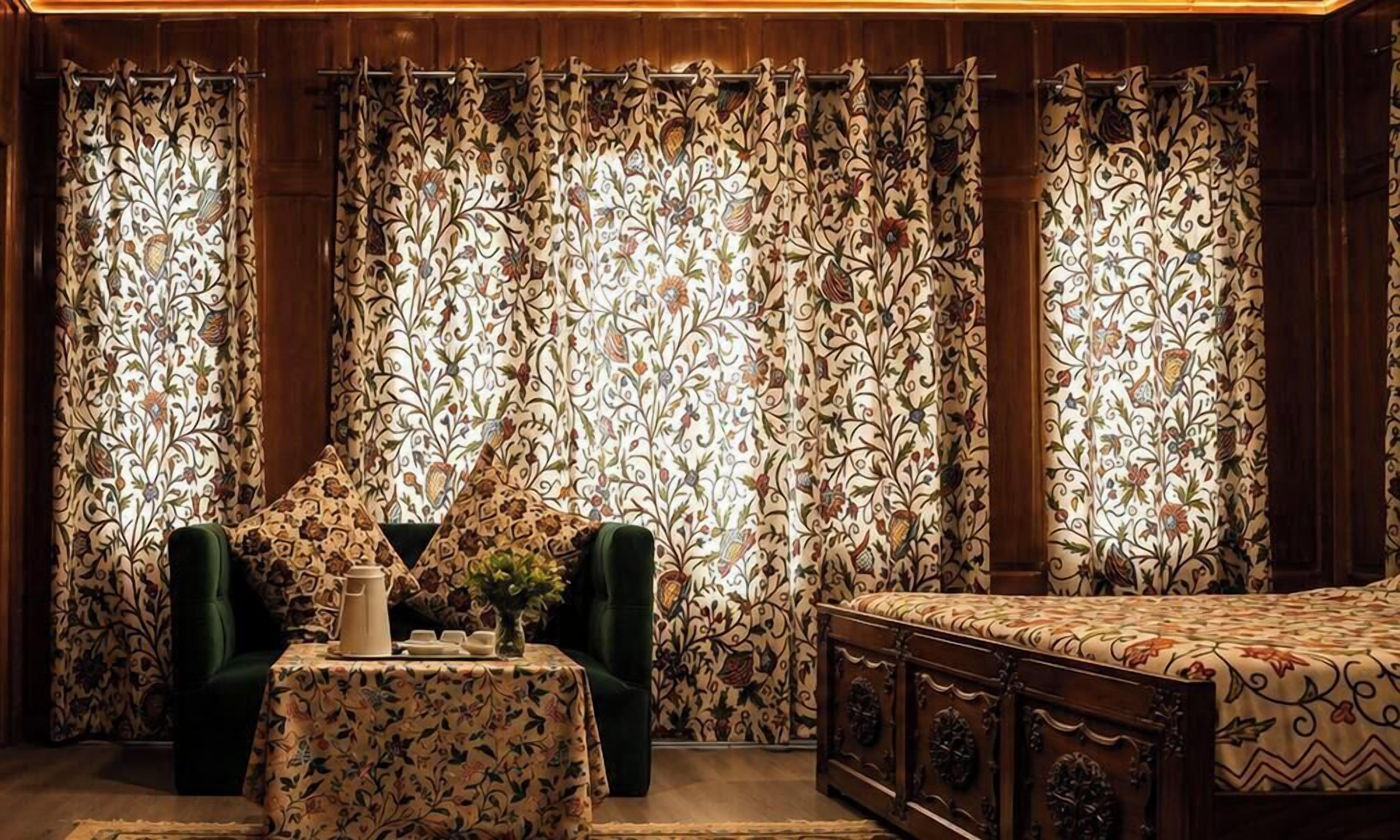 Una habitación con una cama, un sillón verde, una mesita con un jarrón y una jarra, y cortinas con estampado floral.