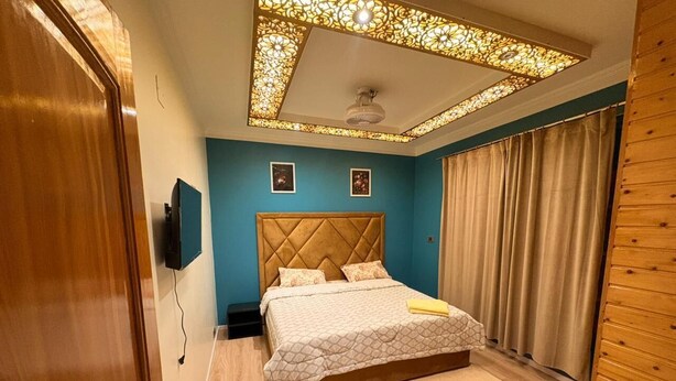 Un dormitorio con cabecera acolchada, cama con colcha estampada, lámpara de techo e televisión montada en la pared.